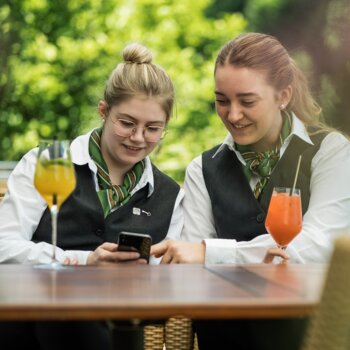 Zwei junge Frauen sitzen im Grünen und zeigen sich den Top Arbeitgeber Hotel Deimann auf Ihrem Smartphone.