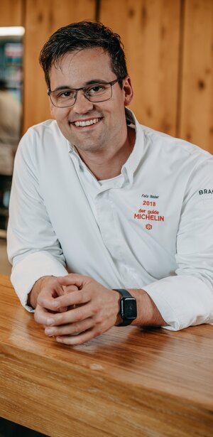 Bild eines lächelnden Mannes in weißer Kochjacke in einer Holzhütte.