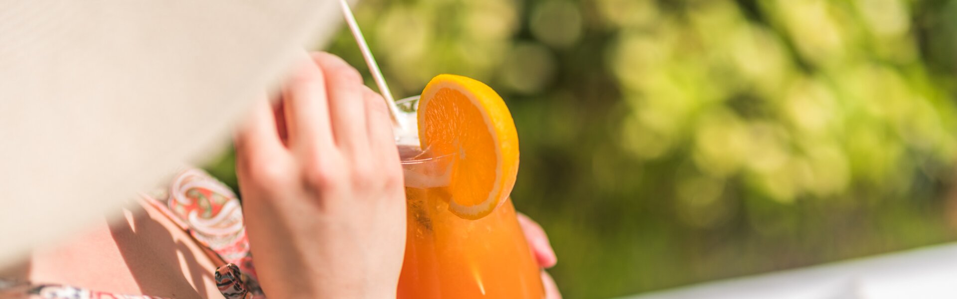 Detailaufnahme eines orangefarbenen Cocktails in der Hand einer Frau, die im Grünen sitzt.