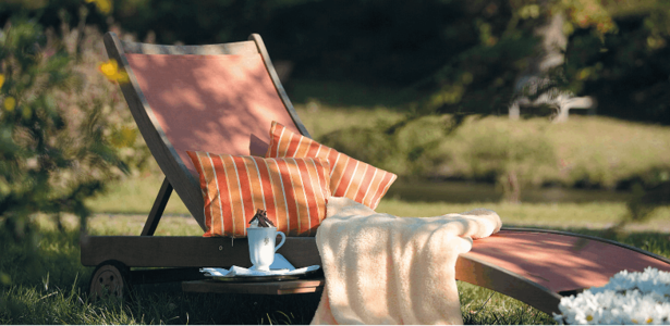 Eine Liege mit Kissen und einer Decke steht in einem Garten, daneben eine heiße Schokolade auf einem Tisch wartend.